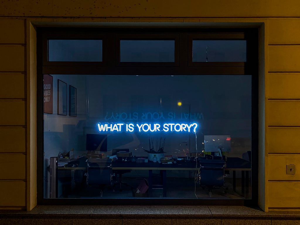 A neon sign reads through an office window: “WHAT IS YOUR STORY?”
