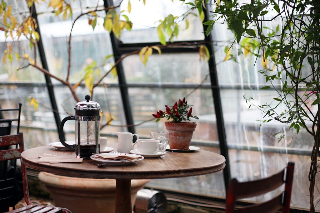 The picture shows a table in cafe with a french press and coffee cups. There are some plants in the background.