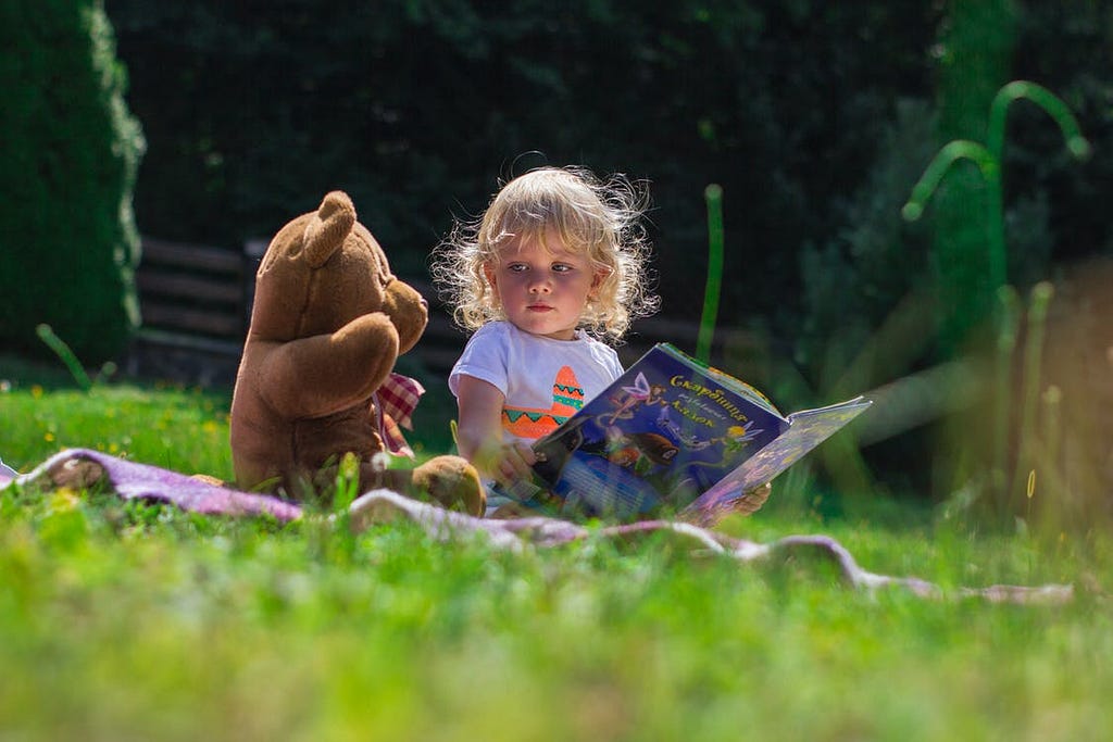 uma foto de uma criança de pele clara e cabelos loiros cacheados e curtos sentada em um pano, que está na grama, ao lado de um urso de pelúcia. a criança segura um livro e olha para o urso de pelúcia.