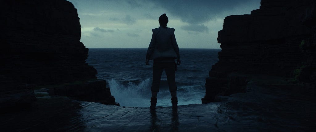 Rey stares out at the crashing waves on Ach-To, facing a grey and cloudy sky.