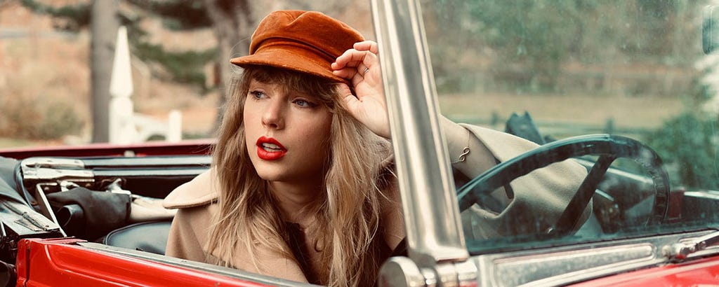 An image of a blonde woman (Taylor Swift) wearing red lipstick sitting in the driver’s seat of a red convertible. She is wearing an orange newsboy cap and a beige coat and her eyes are cast to the side. She has the bill of her cap between her thumb and forefinger. Trees can be seen in the background of the photo.