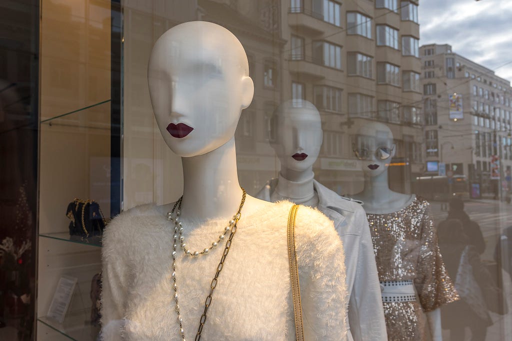 A row of bald white mannequins displaying jewelry in a shop window; apartment buildings are reflected beside them