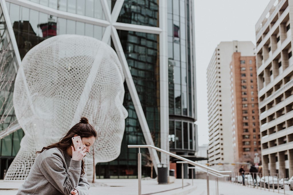 woman sitting outside talking on phone next to futuristic white sculpture of human head