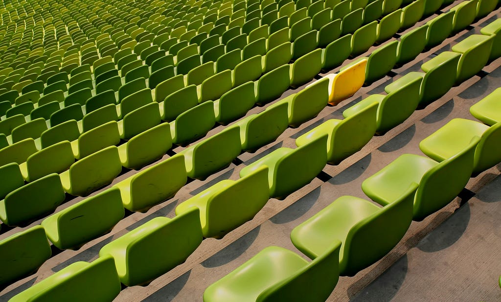 One yellow chair amongst many green chairs in a stadium