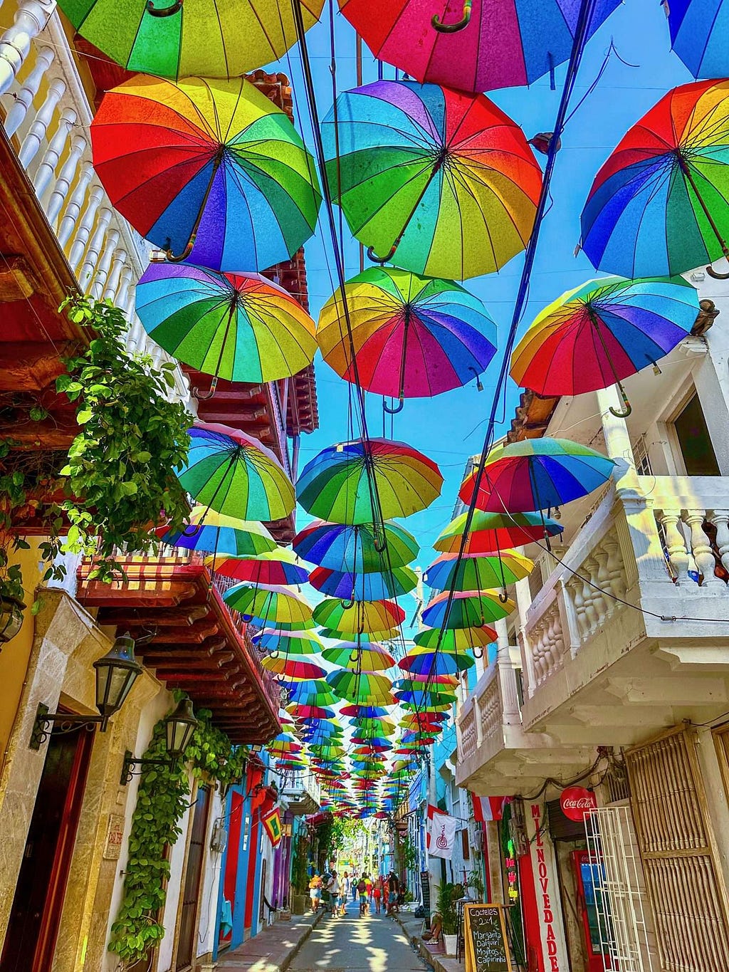 A narrow street in Cartagena, Colombia is adorned with colorful umbrellas suspended above, creating a canopy between the buildings. The blue sky is visible above the umbrellas, and greenery hangs from the balconies.