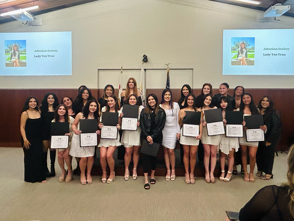 A group picture of the Athenians, dressed in black and white formal attire, the Ladies in the front holding certificates while smiling. A projection of one of the newly initiated Ladies is portrayed on the background wall.