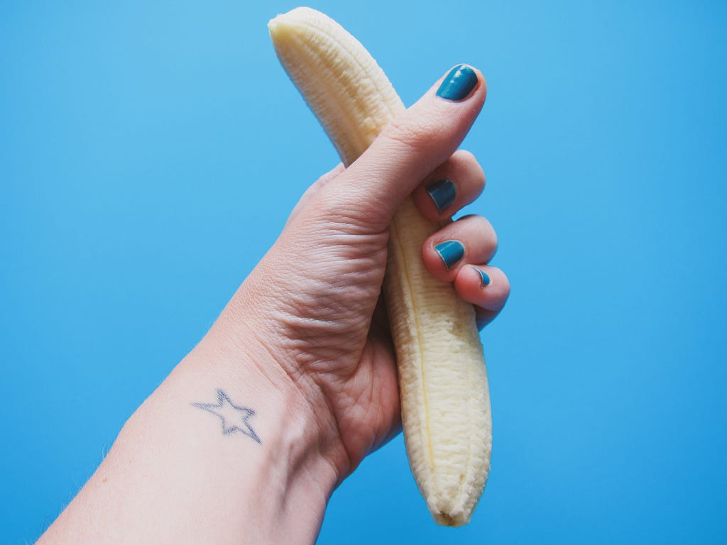 A pink-toned human hand and wrist are visible. There is a hollow star tattoo near the inner wrist. The fingernails are short and painted blue. The hand is clenched around an upright, unpeeled banana. The photo is taken against a blue backdrop.
