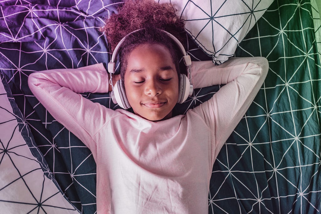 Photo of a middle school girl lying down with her hands behind her head, eyes closed, with headphones on.