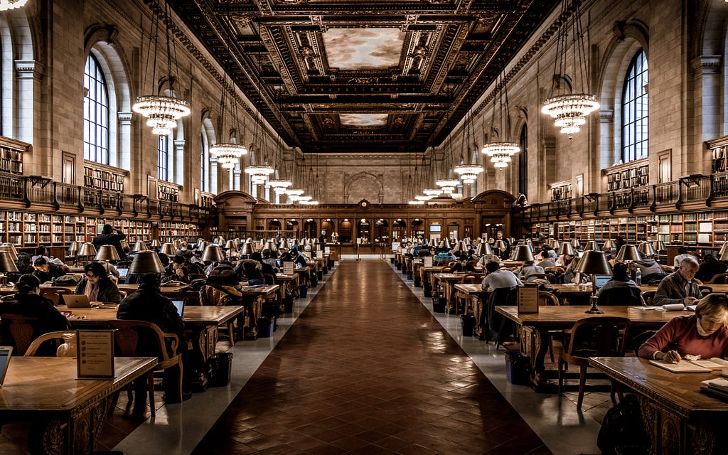 An ornate library with people working at rows of large desks, surrounded by bookshelves and chandeliers.