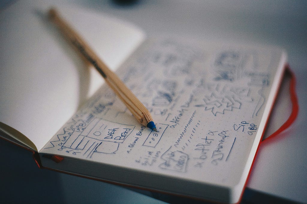 An open notebook lies on a desk, filled with blue-ink sketches, notes, and small diagrams. A wooden mechanical pencil rests diagonally across the page, partially obscuring the drawings. The background is softly blurred, giving the scene a quiet, focused, reflective mood.