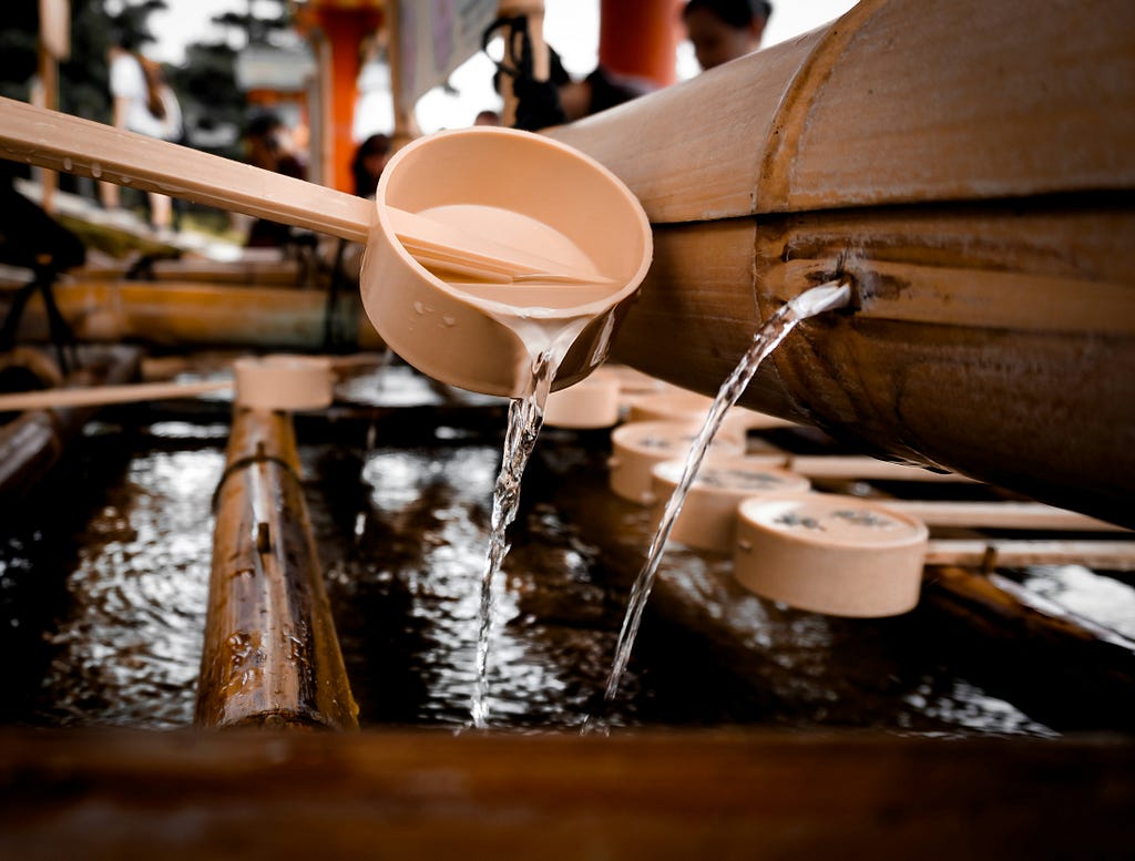 A bamboo ladle pours water into a trough. A bamboo pipe next to it continuously fills the trough with water. More ladles are beneath the pipe.