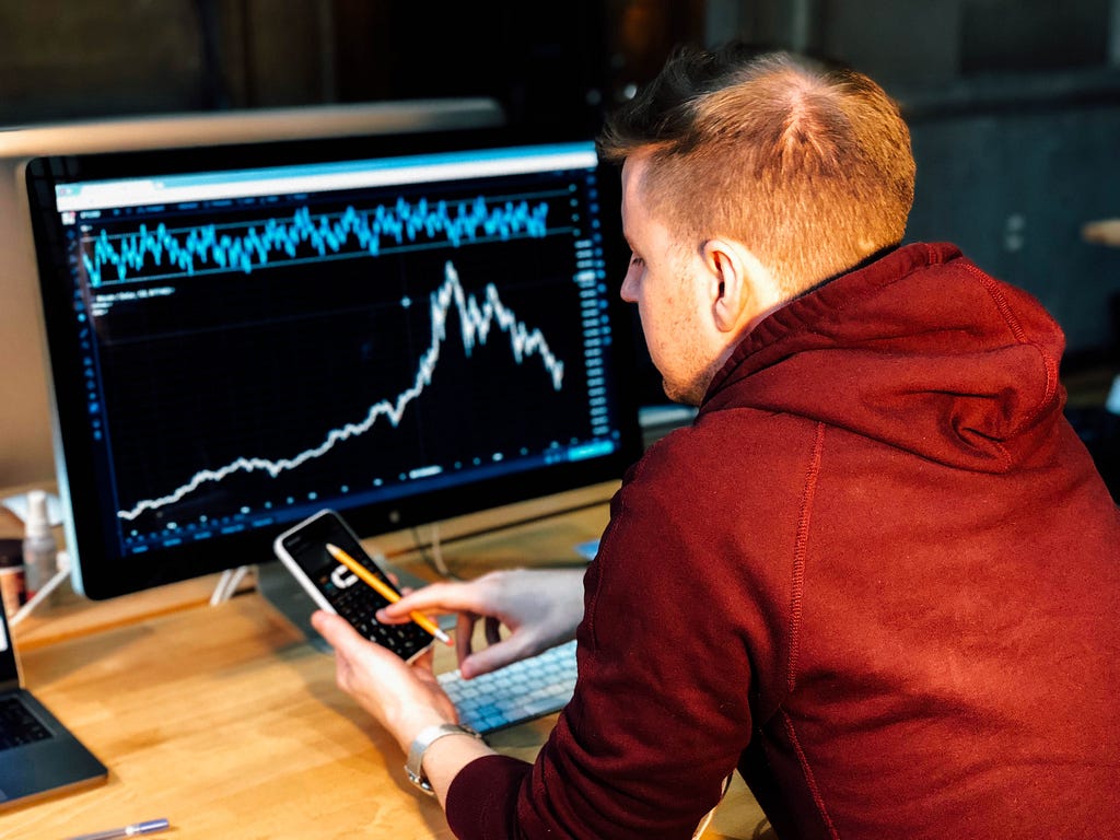 A man who’s using his phone in front a screen with graphs and statistics.