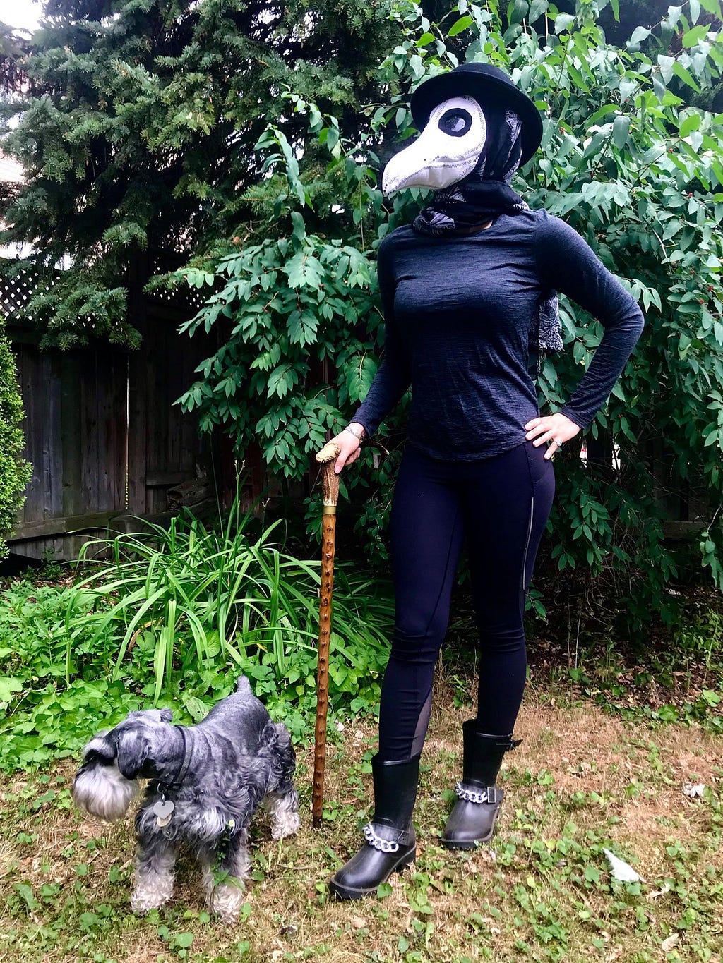 A woman dressed in black and a textile mask stands confidently by her service dog in front of greenery, right hand on cane.