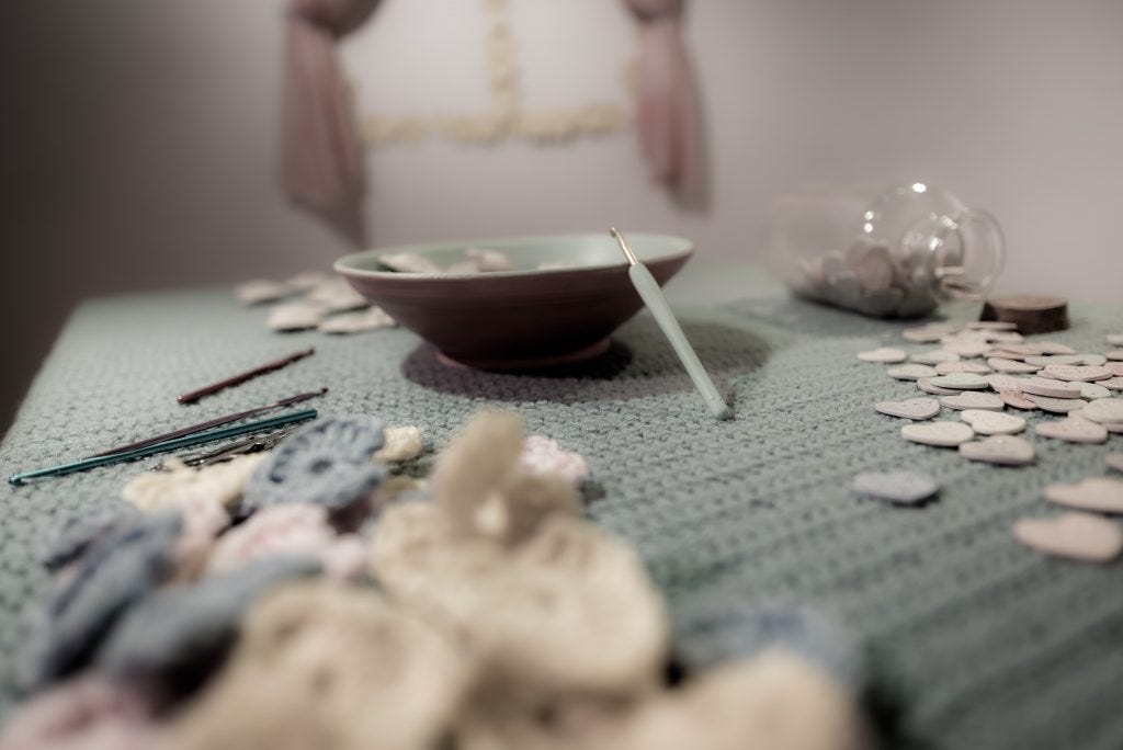 Claudia Solorzano's abstract "kitchen" installation includes a window, an end table with a plant, an alphabet soup of ceramic letters in a corner, and here, in the center of the room, a table and chair, both covered in Solorzano's crochet work. On the table are a bowl of ceramic ravioli candy hearts, a bottle of smaller ceramic candy hearts, crocheting tools, and a pile of small crocheted hearts.