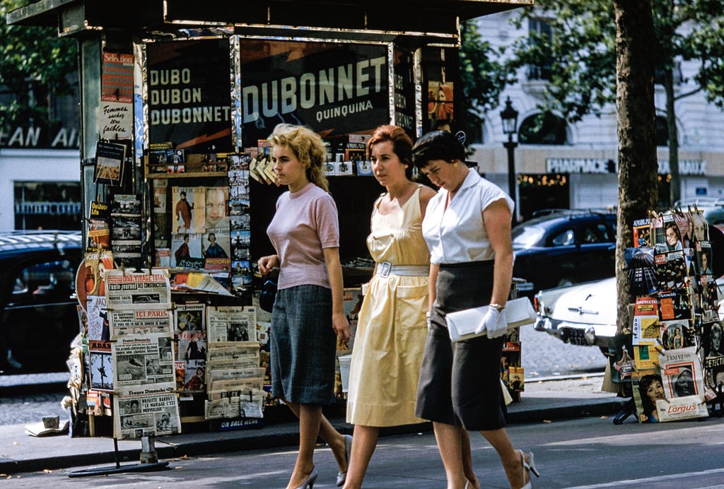 Foto de época de tres mujeres bien vestidas caminando por una calle frente a un puesto de periódicos cubierto de revistas.
