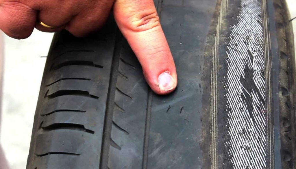 Neumático negro con desgaste extremo. Un lado del neumático está tan desgastado que se observa el alambre metálico. La banda de rodadura ha desaparecido por completo en esta área, dejando expuesta la estructura interna del neumático.