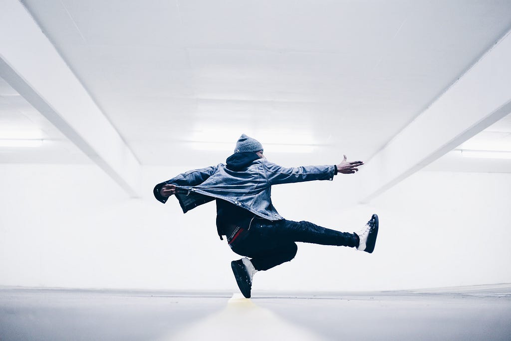 An Unsplash photograph of a young man dancing against a white and gray background. Photo by Drew Dizzy Graham.