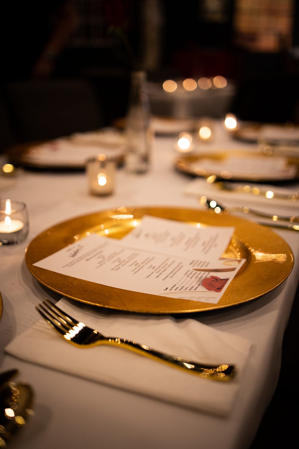A gold dinner plate on a formal dining table with a menu in the foreground is accented by candlelight and out-of-focus dinnerware.