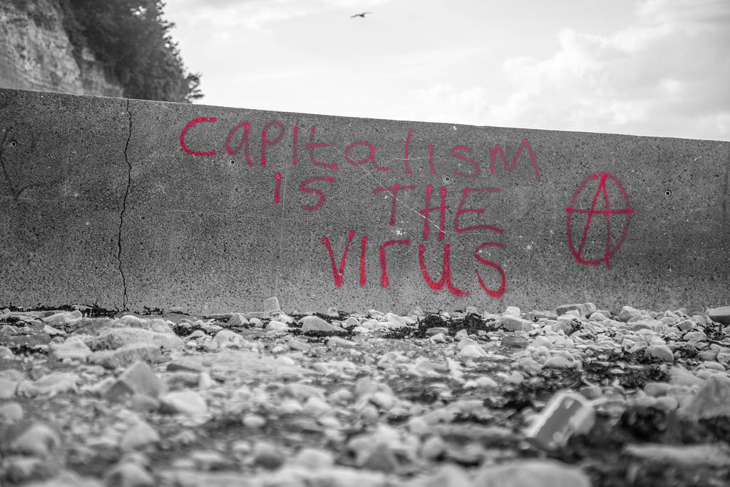 Black and white photo with red letters that say “Capitalism is the virus” with the Anarchy symbol
