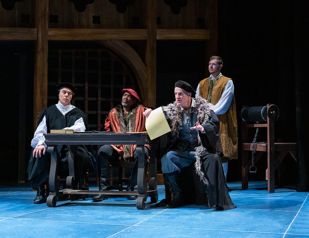 Three men in 16th century robes and hates in black and red sit at a table while the man on the right holds out a piece of parchment paper. A man in a gold robe stands behind them.