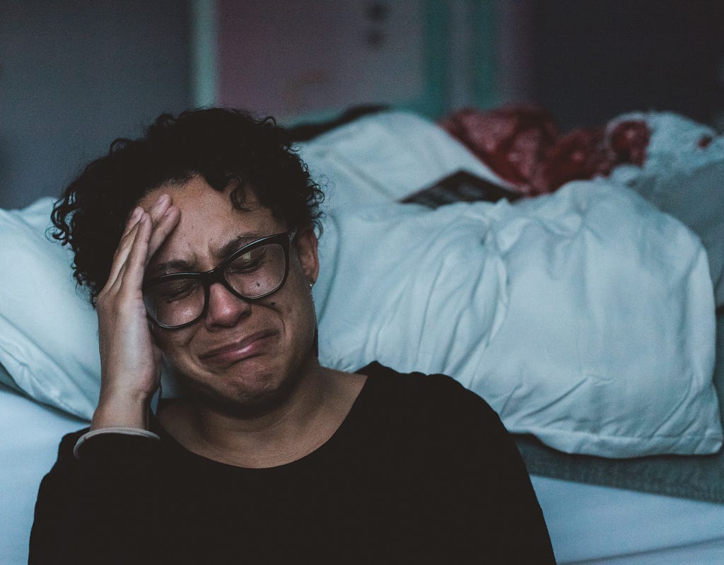 A middle-aged woman sits on the floor beside her bed with her hand to her forehead, crying.