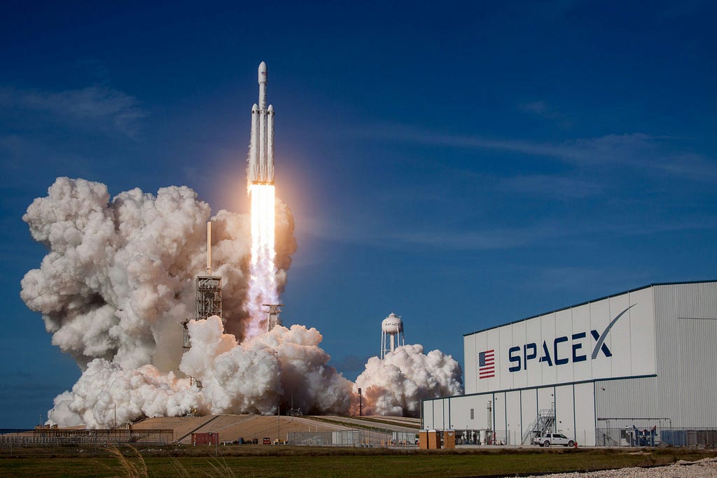 A rocket is taking off and a SpaceX hangar is right next to it.