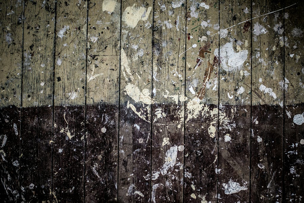 Photo of a wall made of vertical wood boards. The bottom half of the boards are painted black, the top half are beige. There is white and gray paint splattered all over the boards in a very messy way.