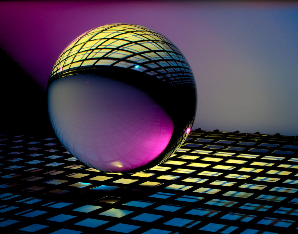 A reflective glass ball placed atop a black grid with lights underneath. The background is a purple-blue, shadowed at the edges. The ball reflects a pinkish color at the base.