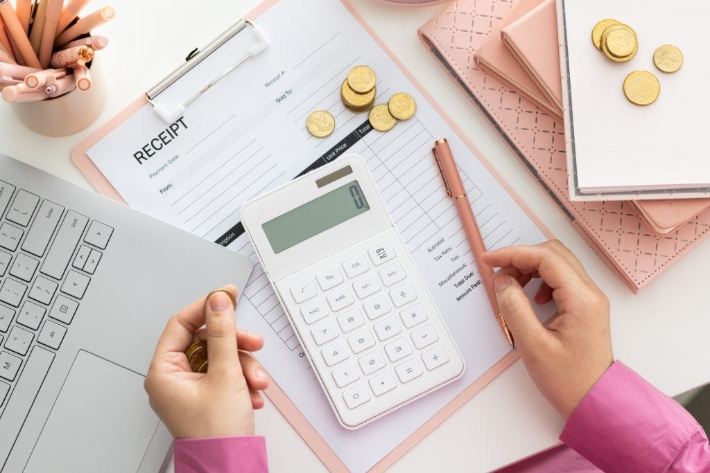 An overhead image of a person in a pink shirt sitting at a desk with a laptop to their left side and a pile of pink books to their right, A white calculator and gold coins scattered about.