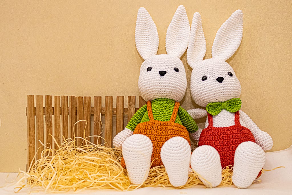 Two crocheted white bunnies in overalls against pastel background