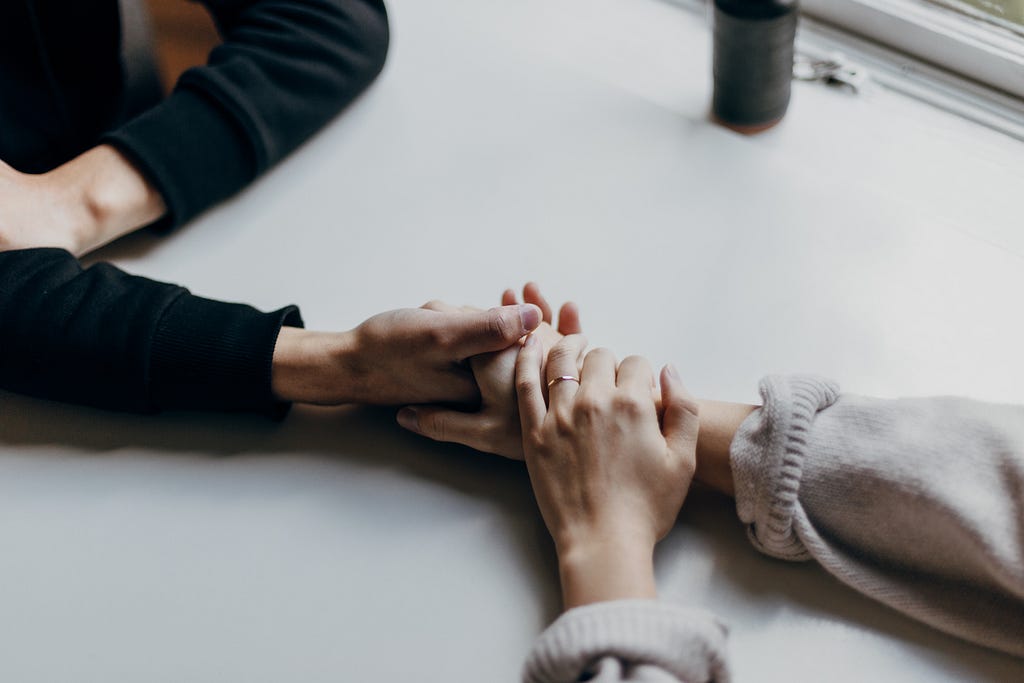 Two people sitting at a table. Closeup of them holding hands, comforting each other.