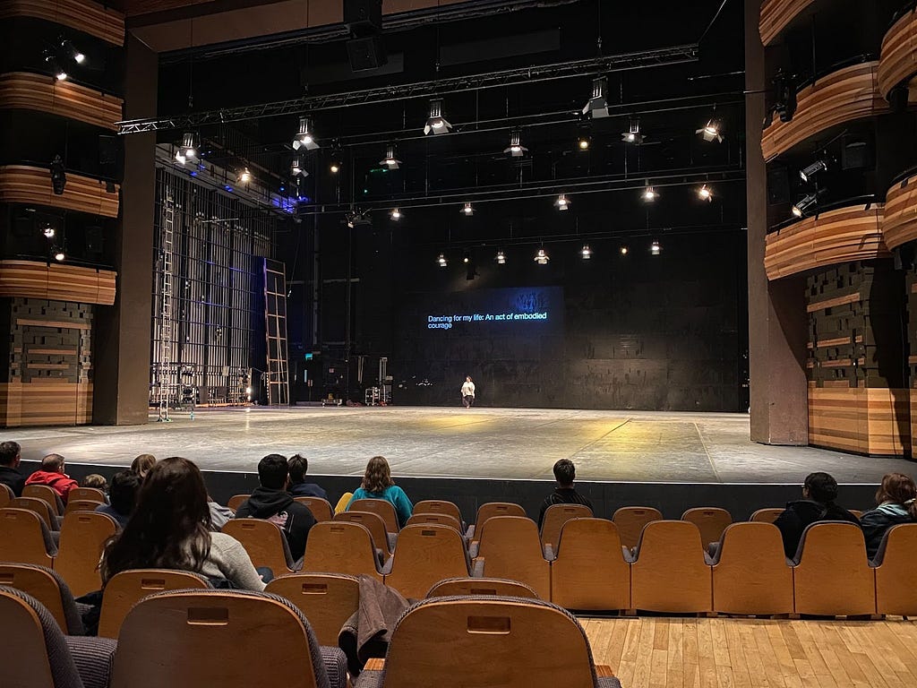 A big stage with not much in it apart from one person standing towards the back of it and a screen reading “Dancing for my life: An act of embodied courage”. Some people sat on brown seats in the audience. House lights are on.