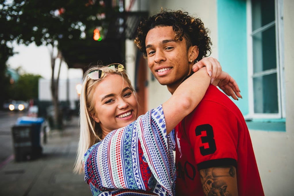 A young woman with straight blonde hair and sunglasses resting on her head drapes her arms around the shoulders of the taller male companion she faces who has dark curls and a bicep tattoo and wears a red soccer jersey.