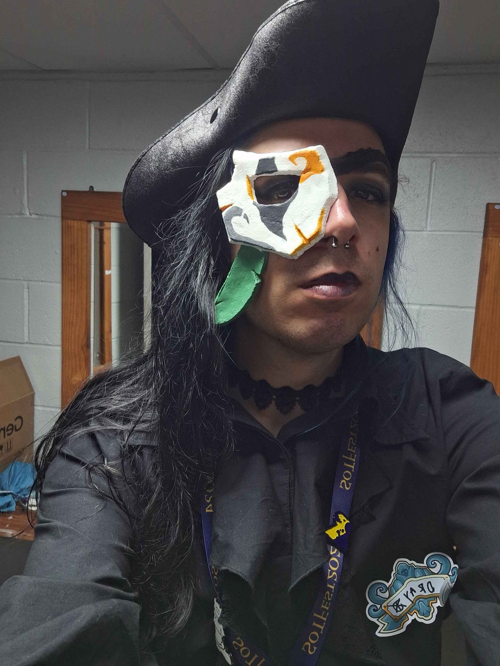 This image is of a single person. They are wearing a black shirt with a black choker. They are wearing dark makeup on their eyes and lips. Covering their right eye is the Daring Deciever Eyepatch, which is a white mask with an eye-hole cut out painted with black and gold accents, it has a green feather accessory attached on the bottom of it. This person is also wearing a tricorn hat.