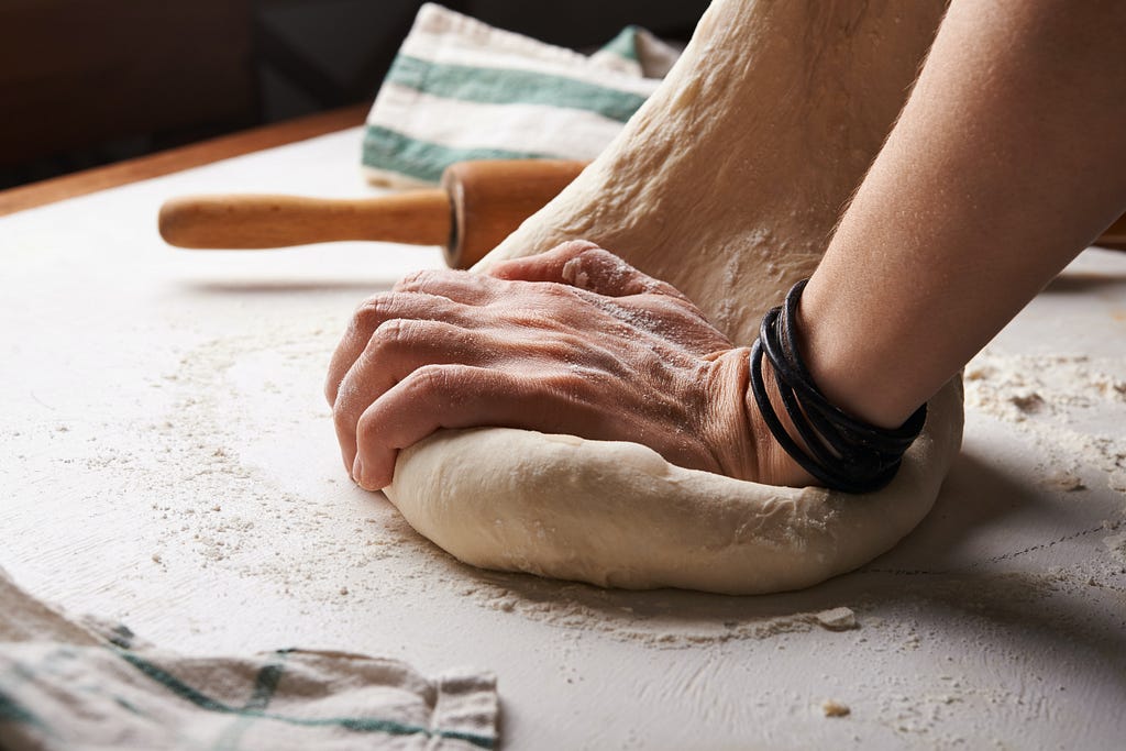 A person’s hand kneading a lump of dough next to a brown rolling pin.