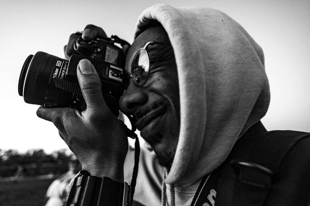 Man wearing a hoodie and glasses, holding camera up to his eye in black and white