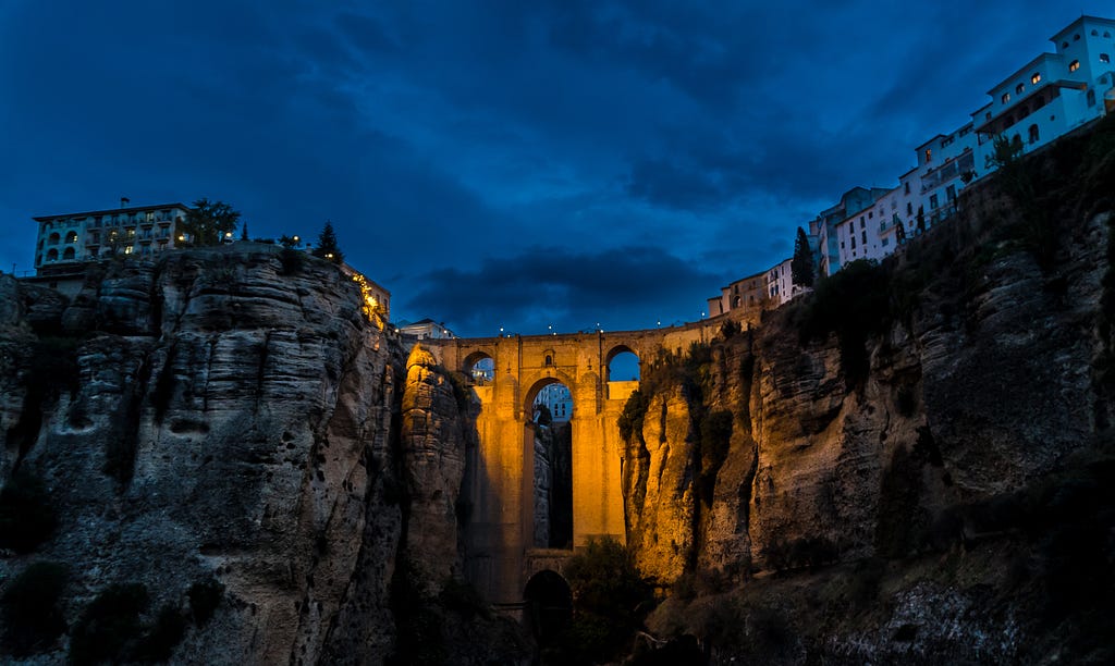A Roman bridge crossing a gorge, lit by dramatic lights.