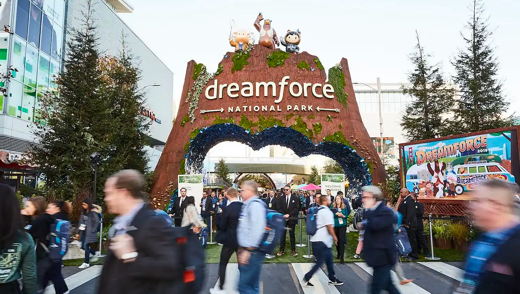 A large structure shaped like a canyon wall that represents the Dreamforce National Park entrance. There’s a cloud-shaped arch cutout. There are lots of people walking through the entrance and also crossing the street in the foreground.