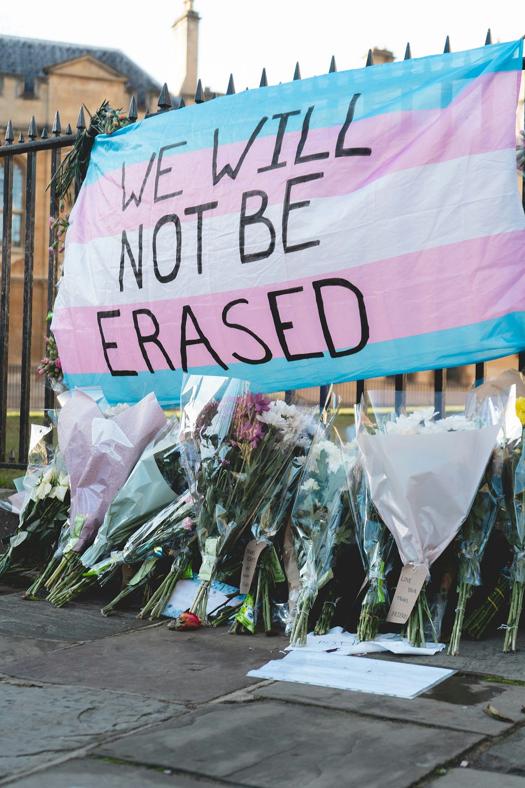 Trans flag on a fence with flowers beneath, likely a memorial. Writing on the flag reads “we will not be erased.”