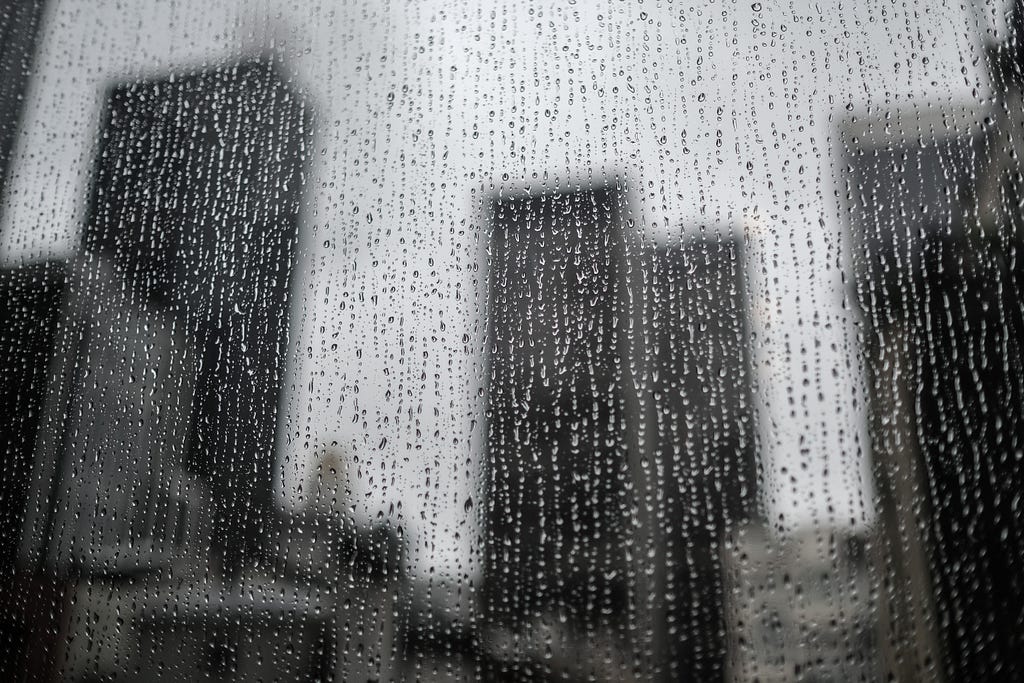 Photo looking out through a window covered with rain drops. There are blurry skyscrapers in the background.