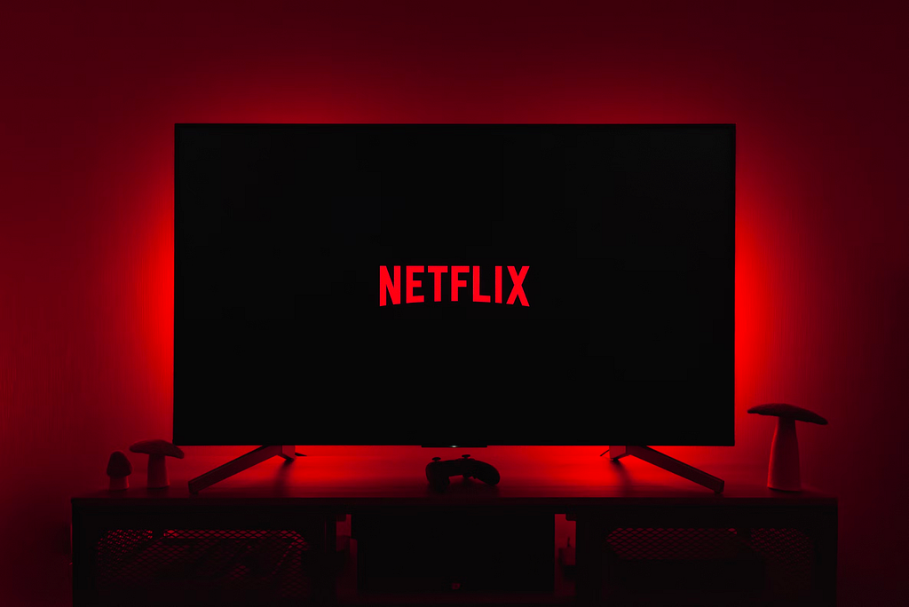 In a dark room there is television with the Netflix logo on screen. There is a ominous red light behind the television.