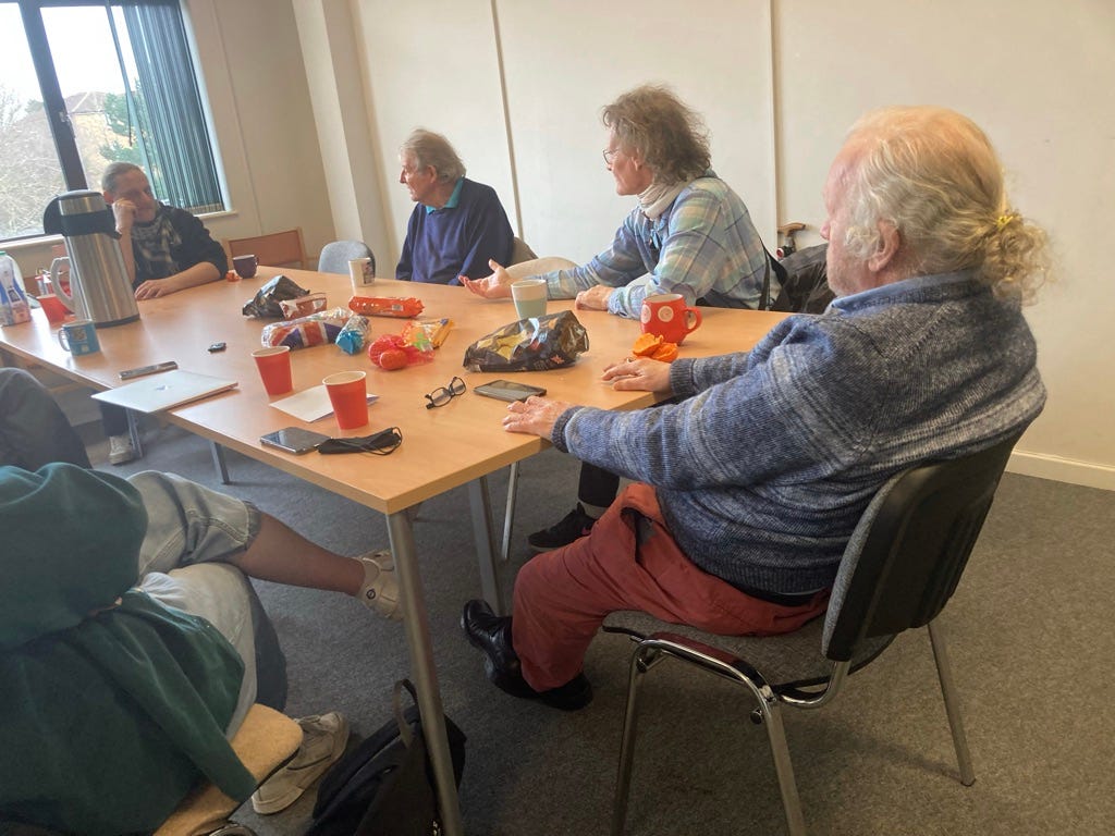 Five people sat around a table with mugs, a coffee dispenser croissants and other bits and bobs on the table.