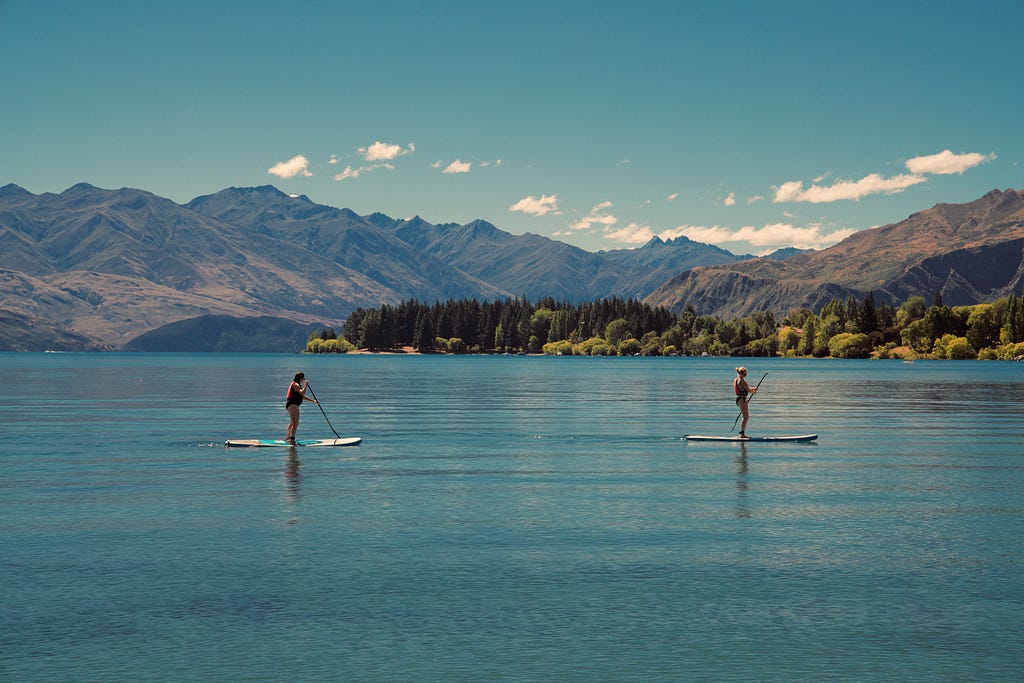 Two people stand-up paddleboard on a lake with mountains in the background.
