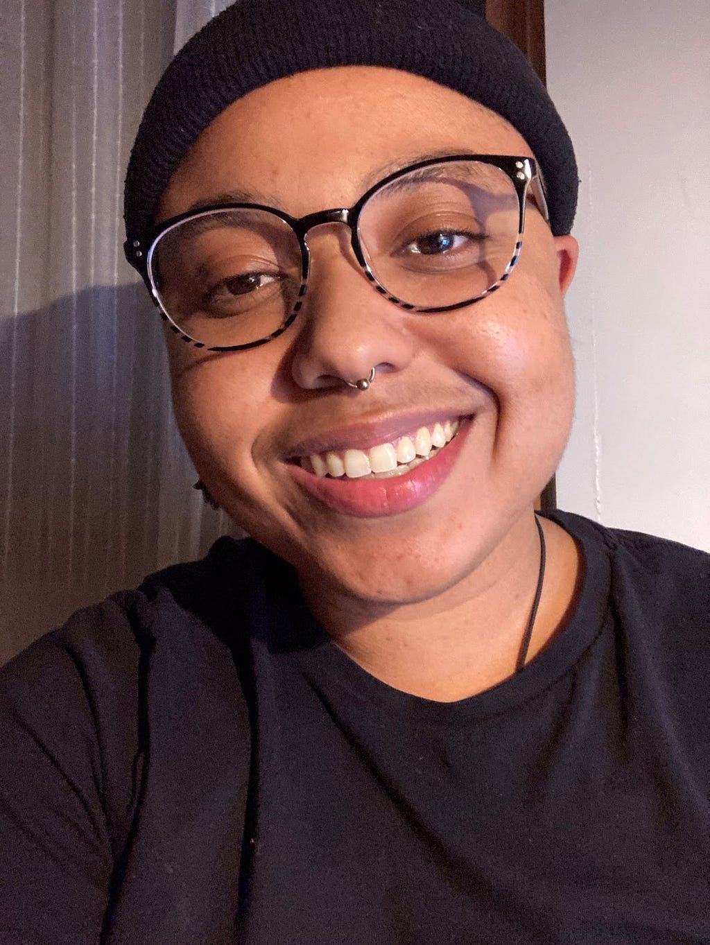 Aiyo smiling and sitting against a white background near a window covered by a white lace curtain to their right. They are wearing a black beanie, black t-shirt, and plastic glasses with a black frame — there are black and clear stripes on their glasses. They have a septum piercing and are looking at the camera.