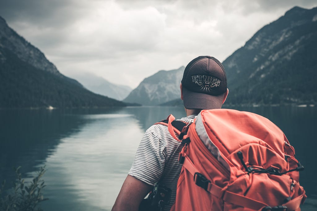 A man with a hiking backpack stares at a lake surrounded by mountains