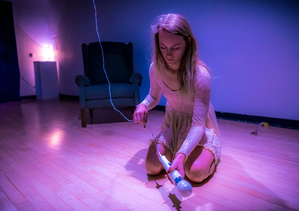 Performance Artwork by Cortnee Brush. Brush is wearing a white, lace dress, and is kneeling on the gallery floor. She holds a vibrator in her hand. The vibrator has a cord that goes up to the gallery ceiling.