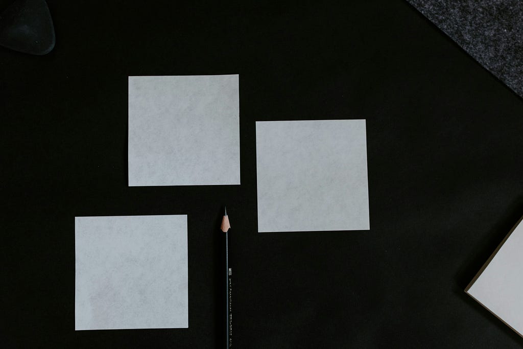 Three blank square sticky notes arranged loosely on a dark surface with a sharpened black pencil placed below them. The scene is minimal and flat-lay, suggesting planning or organizing. No text or markings on the notes.