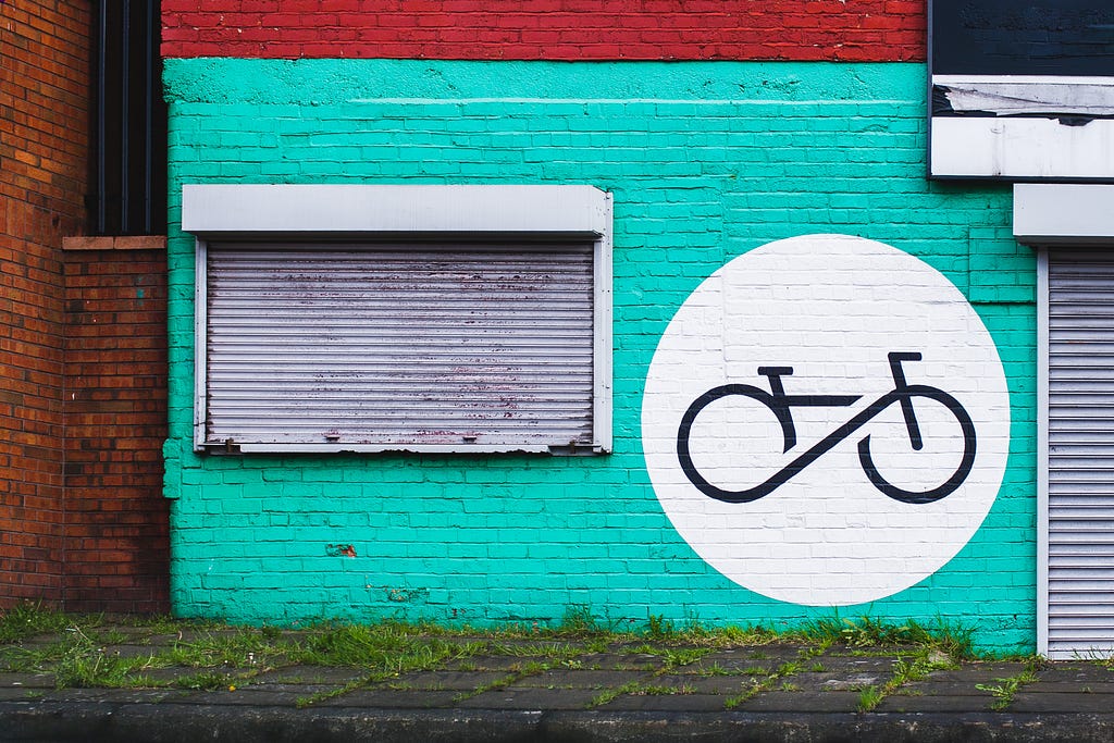 A turquoise brick building with closed shutters and a bike painted on the wall