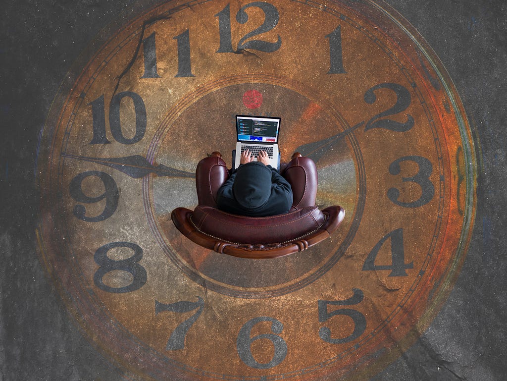 A wooden floorboard designed as a large clock. A person working on a laptop. They’re sat in an armchair. The armchair is positioned in the middle of the big clock, viewed from above.