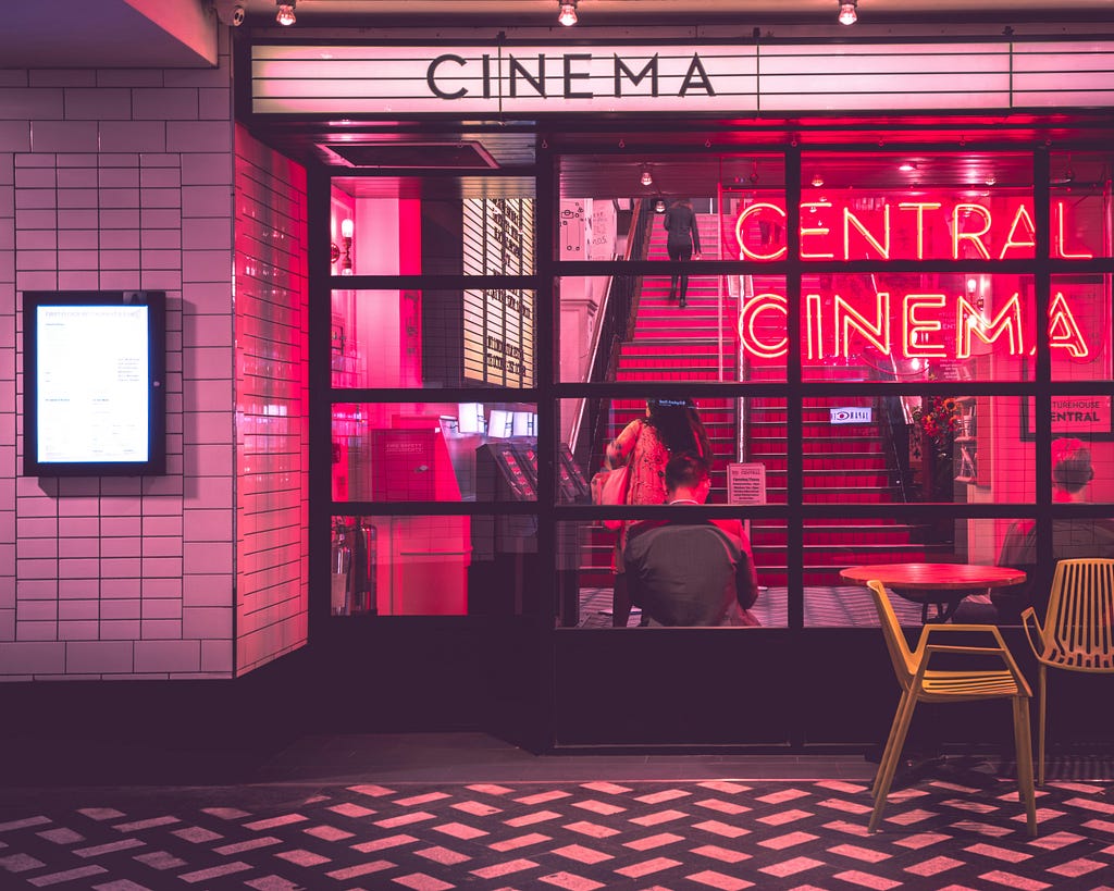 An image shows a wall with glass panels that reveals what’s going on inside the building. Inside, there are stairs leading up. A neon letter light spells out the words “Central Cinema” and hangs just near the glass panels. A couple of yellow chair are outside the panel.